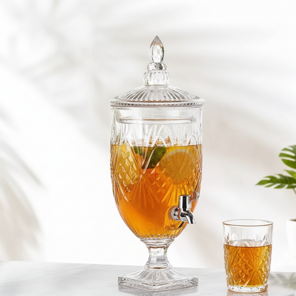 Glass beverage dispenser with lemon slices and a small glass of tea on a marble surface.